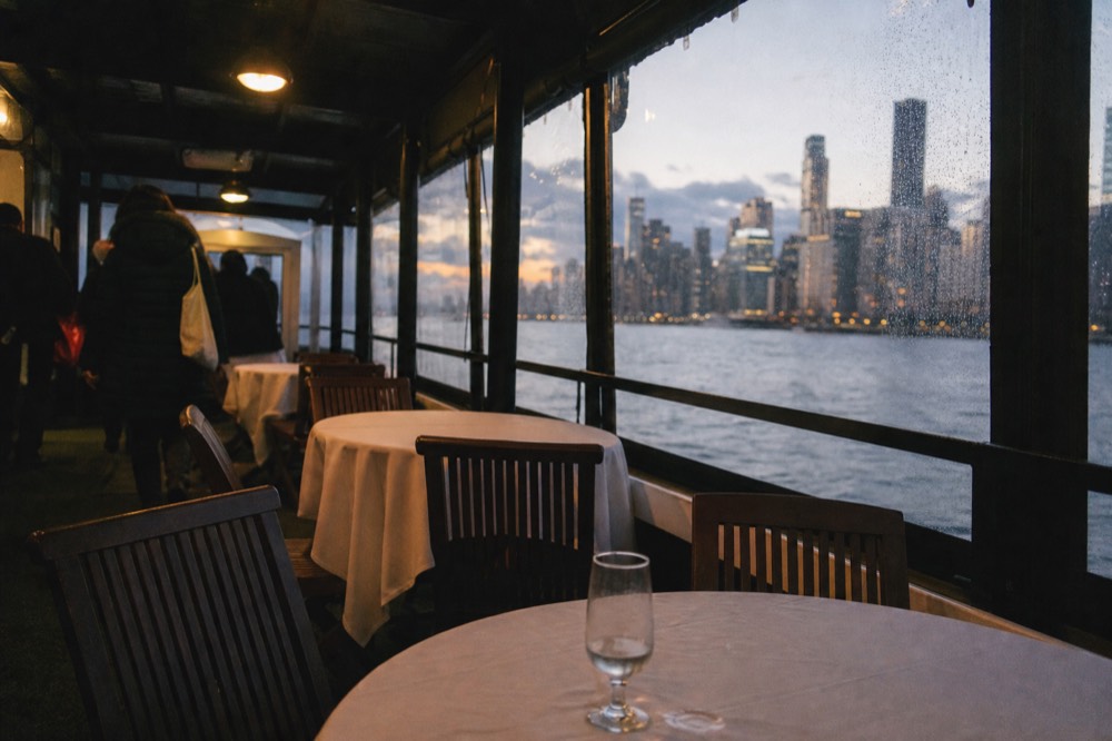 A riverboat dining deck set for dinner with the Savannah skyline through the windows, no text, no logos, no people