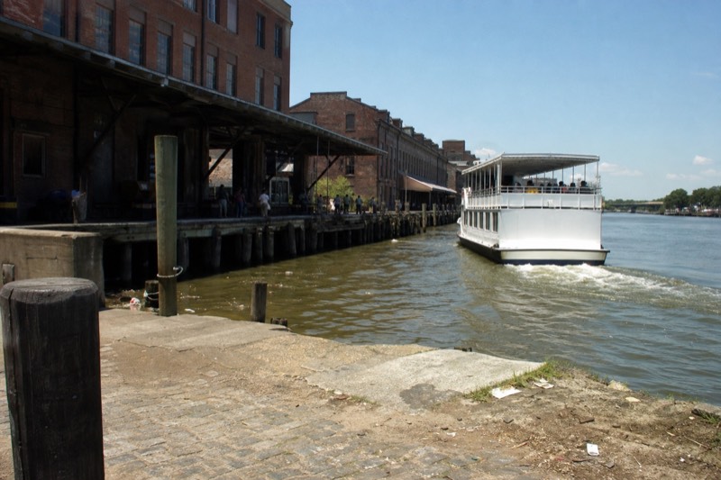 A narrated riverboat easing past Savannah's historic brick riverfront warehouses on River Street under midday light
