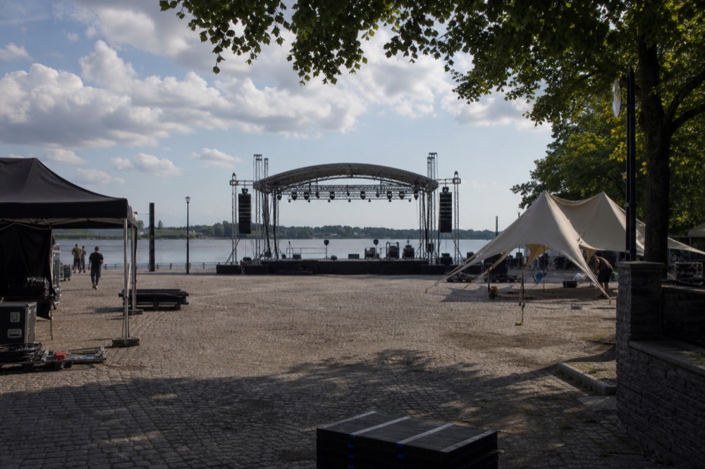 Festival crowd area on a riverfront plaza with a stage and the river behind, no text, no logos, no people