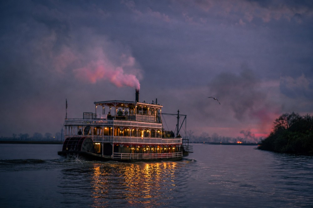 A paddlewheel riverboat glowing with warm interior lights against a dusk sky, no text, no logos, no people