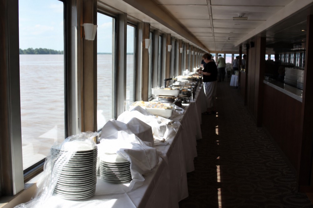 A riverboat dining deck set with buffet tables beside wide windows overlooking the river, no text, no logos, no people