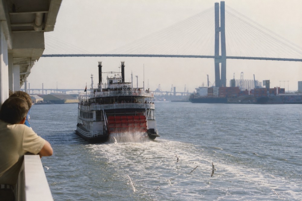 A large paddlewheel riverboat cruising up the Savannah River with a container port and bridge in the distance, no text, no logos, no people