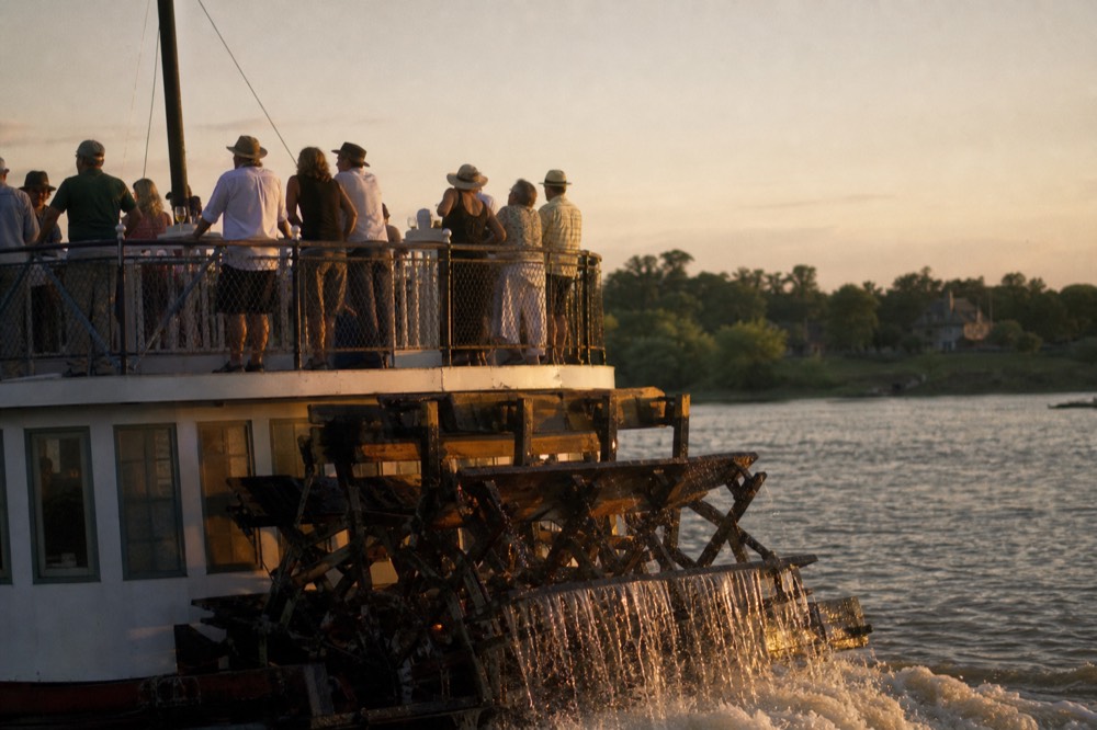 A large paddlewheel riverboat with passengers on the open upper deck during golden hour, no text, no logos, no people