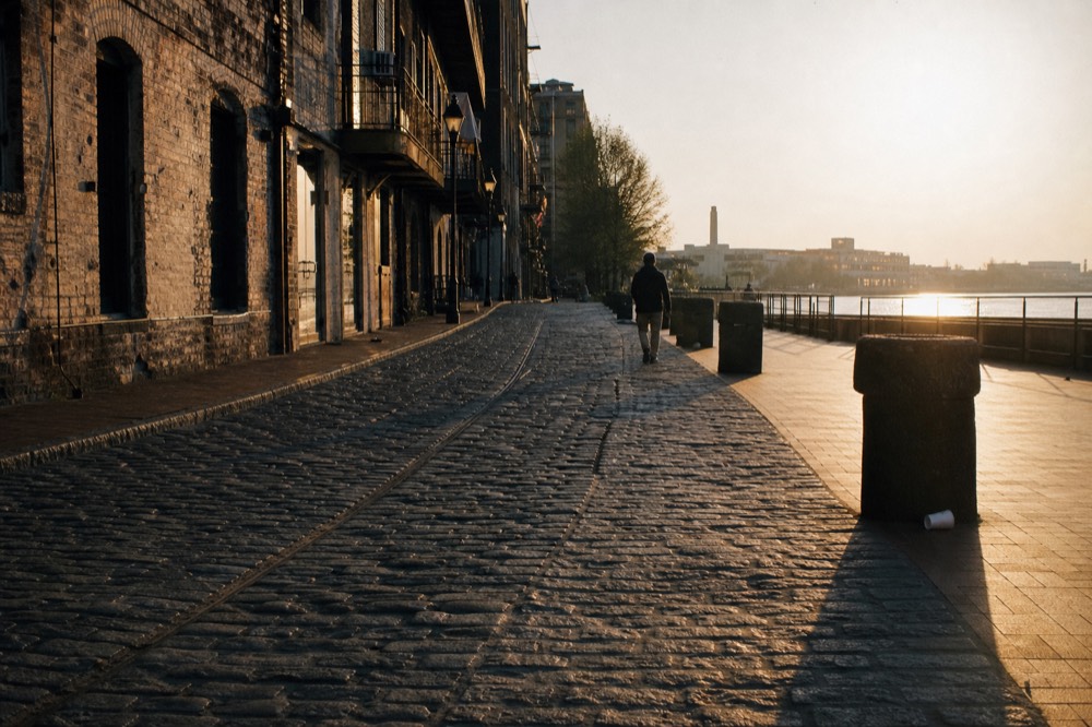 Golden hour light on River Street cobblestones and brick warehouses with the river beyond, no text, no logos, no people
