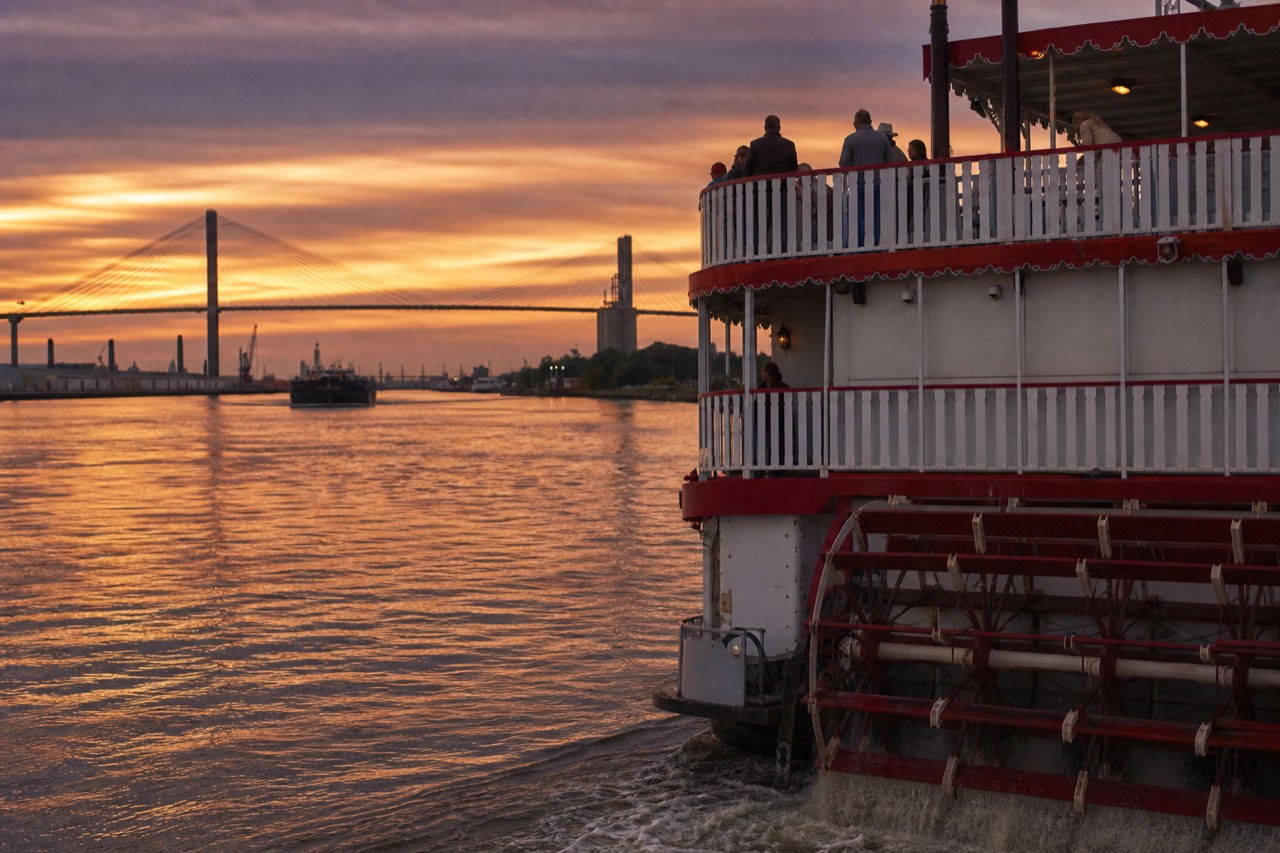A red-and-white paddlewheel riverboat on the Savannah River at golden hour with the Talmadge Bridge in the background, no text, no logos, no people