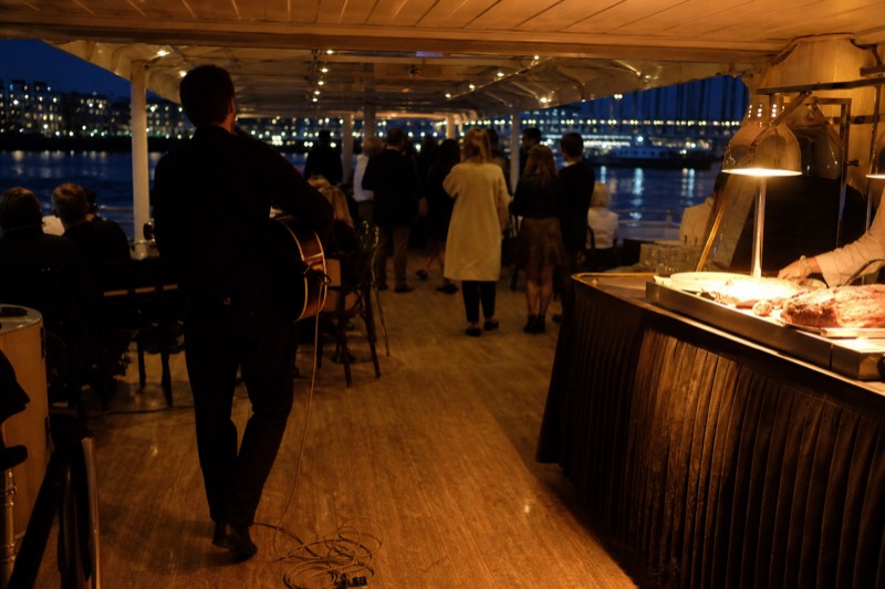 Dinner tables set on the covered deck of a Savannah riverboat at dusk, city lights reflecting on the Savannah River, departing from the historic River Street waterfront in downtown Savannah