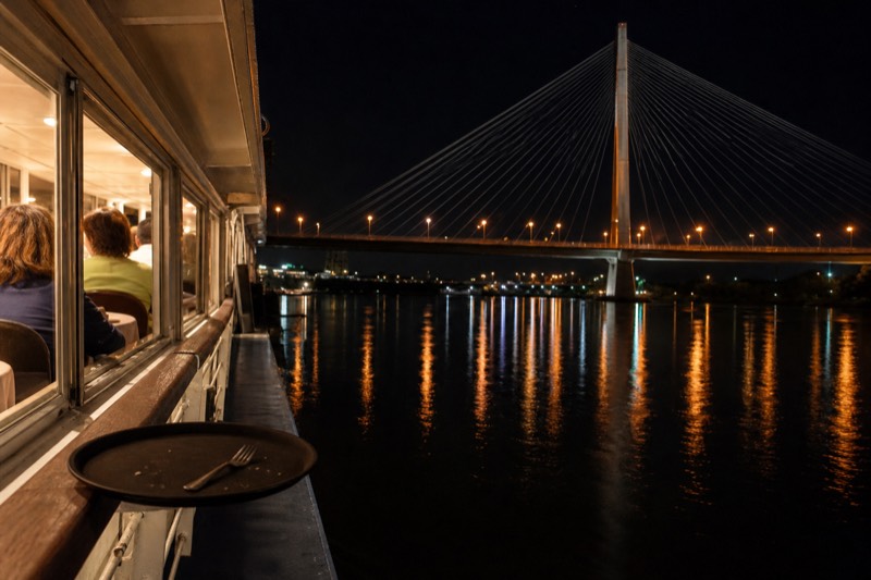 Dinner tables set on the covered deck of a Savannah riverboat at dusk, city lights reflecting on the Savannah River, departing from the historic River Street waterfront in downtown Savannah