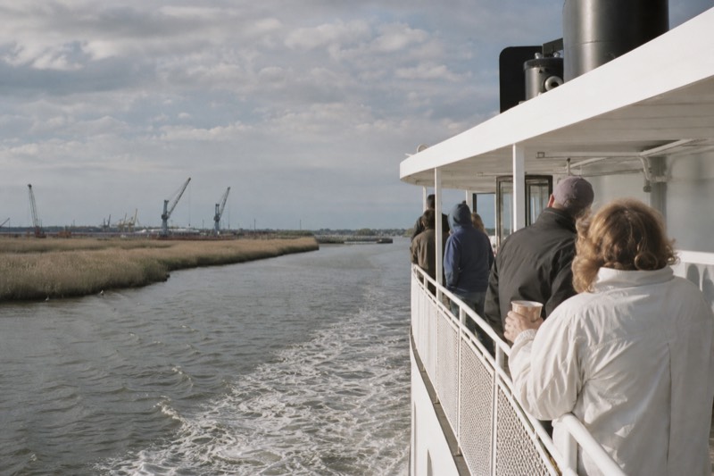 A narrated riverboat easing past Savannah's historic brick riverfront warehouses on River Street under midday light, departing from the historic River Street waterfront in downtown Savannah