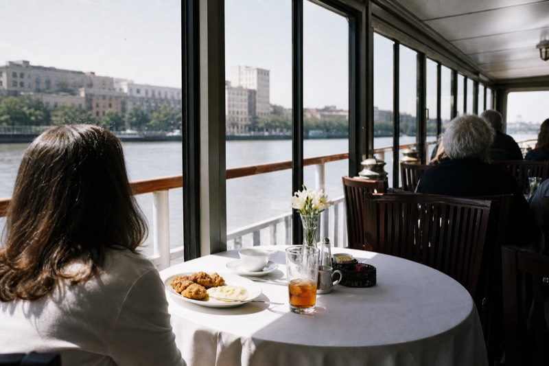 A narrated riverboat easing past Savannah's historic brick riverfront warehouses on River Street under midday light, departing from the historic River Street waterfront in downtown Savannah
